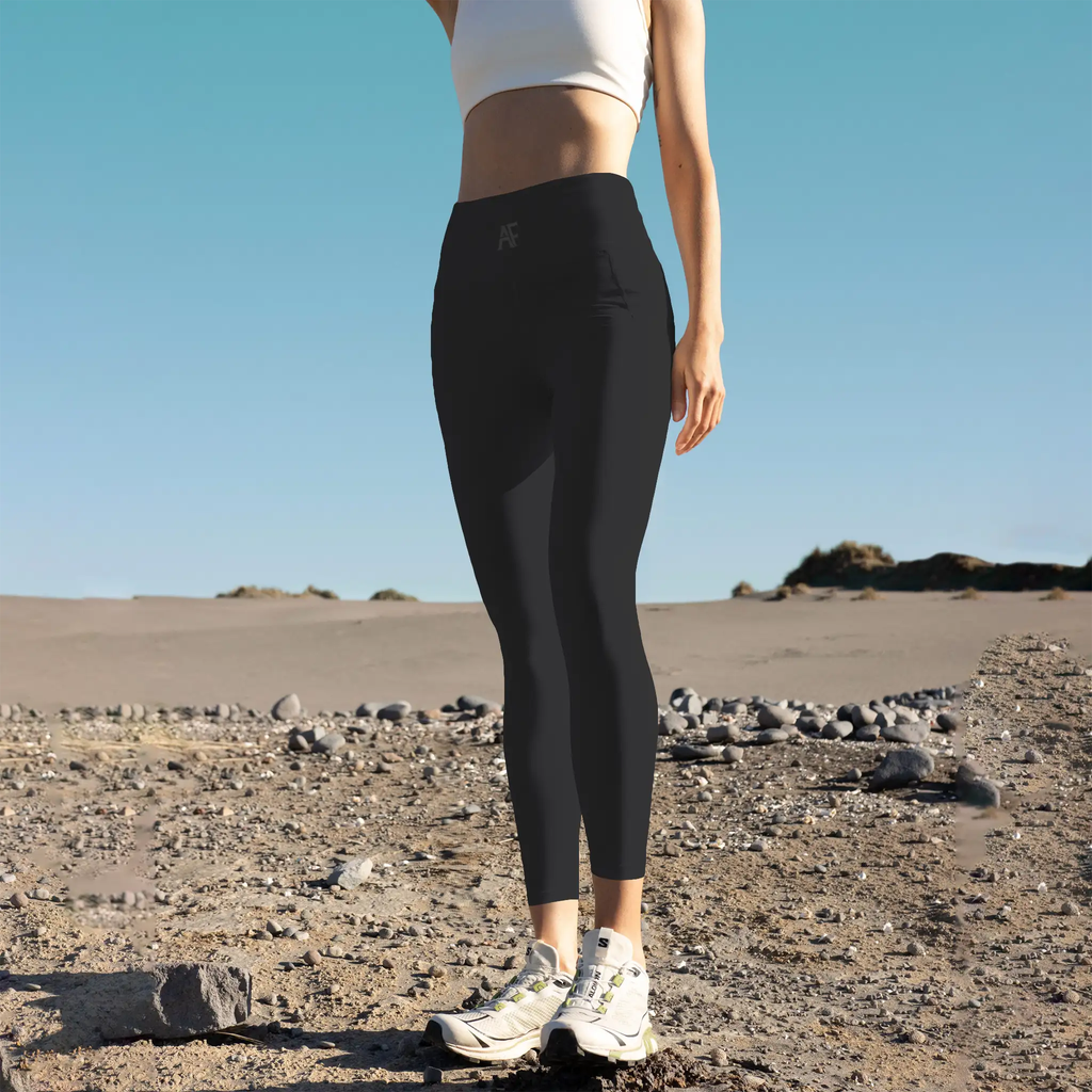 A person wearing black Aphrodite leggings and white sneakers is standing on a rocky beach.